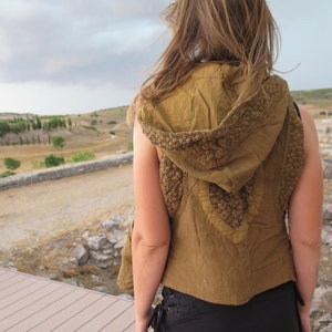 May include: A sleeveless, olive-green hooded vest with a textured, decorative trim around the hood and shoulders. The vest is made of a soft fabric and is worn over a black top. The background features a scenic view of a landscape with ancient ruins.