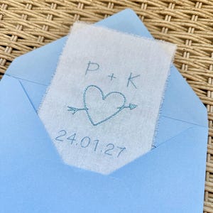 May include: A light blue paper envelope containing a rectangular piece of off-white fabric with frayed edges. The fabric features blue embroidery of the letters P + K, a heart pierced by an arrow, and the date 24.01.27 stitched below the heart.