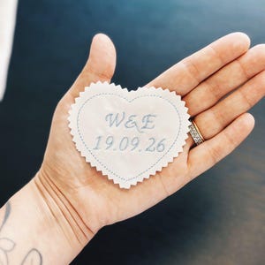 Puede incluir: Un parche de tela en forma de corazón con bordes dentados, que presenta un bordado en azul claro con las letras W&E y la fecha 19.09.26. El parche tiene un borde cosido en azul claro que sigue la forma del corazón sobre una base de tela crema.