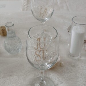 May include: Two clear glass wine glasses featuring a white lace doily appliqué on the bowl. The front glass displays the gold-coloured text "JUST" while the glass positioned behind it displays the gold-coloured text "ST" in a stacked arrangement.
