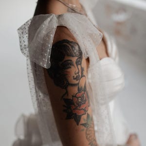 May include: A white tulle dress with polka dots and a ruffled strap. The dress is worn by a person with a tattoo of a woman's face and a rose on their arm.