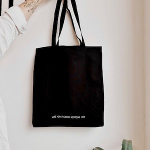 May include: A black tote bag with two shoulder straps, featuring the white text ARE YOU FUCKING KIDDING ME? printed in a small, simple font near the bottom centre of the bag. The bag is made of a solid black fabric and has a rectangular shape.