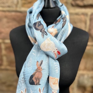May include: A light blue scarf featuring a repeating pattern of various rabbits in brown, white, and gray. The scarf has a fringed edge and is draped over a black mannequin. The background is a brick wall.