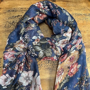 Può includere: Una sciarpa blu navy con un motivo floreale. La sciarpa presenta grandi fiori nei toni del bianco, rosa, arancione e giallo, su uno sfondo blu scuro. La sciarpa è realizzata in un materiale leggero.