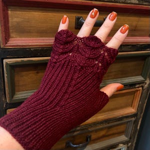 Può includere: Un guanto senza dita bordeaux con un motivo a conchiglia sul dorso della mano. Il guanto è lavorato a maglia con un motivo a coste sul polso e sul palmo. Lo sfondo è una cassettiera in legno con più cassetti.