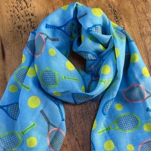 May include: Light blue scarf with a tennis theme. The scarf features a repeating pattern of tennis rackets and yellow tennis balls. The rackets have green, pink, and blue accents. The scarf is made of a lightweight fabric.