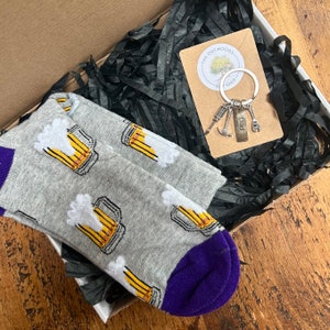 May include: A gift box filled with black shredded paper. The box contains a pair of gray socks with a beer mug pattern and a silver keychain with a small hammer, screwdriver, wrench, and a tag that says "Dad".