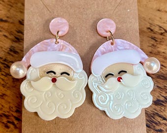 May include: A pair of pink and white Santa Claus earrings with a pearl accent. The earrings are made of resin and have a whimsical design.