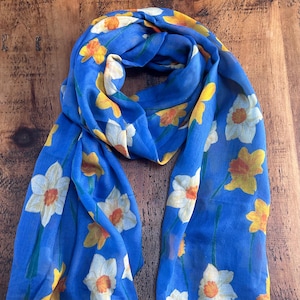 May include: A blue scarf with a floral pattern of white, yellow, and orange daffodils. The scarf is made of a lightweight fabric and is draped on a wooden surface. The flowers are evenly spaced across the scarf.