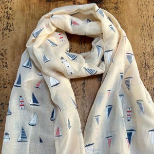 May include: A cream-coloured scarf with a nautical design. The scarf displays a repeating pattern of sailboats, lighthouses, and nautical flags in navy blue, red, and white. The scarf is made of a lightweight fabric.