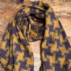 May include: Mustard yellow and dark purple scarf with a geometric pattern. The scarf is made of a soft material and is draped on a wooden surface. The scarf has a fringed edge.