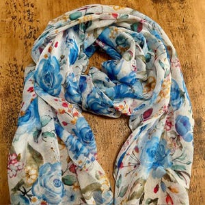 Può includere: Una sciarpa bianca e trasparente con un motivo floreale blu. La sciarpa presenta un design ad acquerello con rose blu e altri fiori.