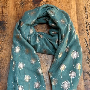 May include: A teal blue scarf with a gold dandelion print. The scarf is folded and displayed on a wooden surface.