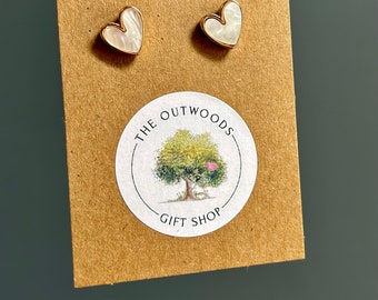 Pode incluir: Um par de brincos de botão em forma de coração com uma borda dourada e um centro branco perolado. Os brincos são exibidos em um suporte de papelão marrom com um adesivo circular que diz "THE OUTWOODS GIFT SHOP" em torno de uma ilustração de árvore.