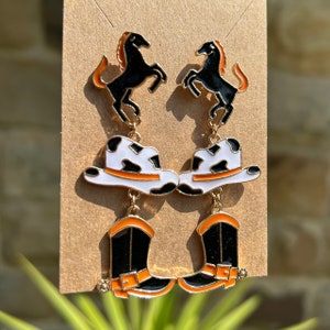 May include: A pair of dangle earrings featuring a black horse, a white cowboy hat with black spots and an orange band, and a black cowboy boot with an orange top. The earrings are gold-toned.