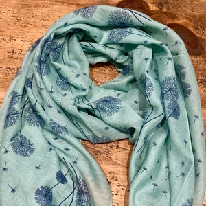 May include: A light teal scarf with a repeating pattern of dark blue dandelion seed heads and floating seeds. The scarf is draped, showing the soft, flowing fabric.