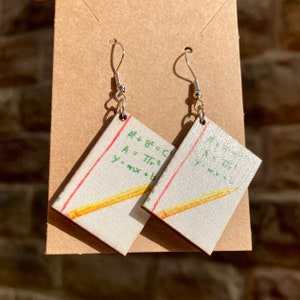 May include: A pair of wooden earrings shaped like a notebook page with a pencil drawn on it. The notebook page has math equations written on it in green ink. The earrings are white with a light brown wood grain.