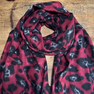 May include: A burgundy scarf featuring a black and grey leopard print design. The scarf is made of a soft material and is draped on a wooden surface. A fashion accessory.