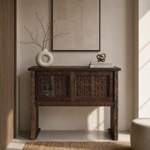 May include: A dark brown wooden console table with two cabinet doors featuring intricate carved geometric patterns and a circular metal pull handle. The table surface holds a white ceramic donut-shaped vase with thin branches and two stacked books.
