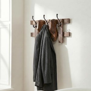 May include: A wall-mounted coat rack made of horizontal wooden planks featuring three dark metal hooks and two vintage wooden shoe lasts attached to the center. A charcoal gray wool coat hangs from the center hook, covering the lower portion of the rack.