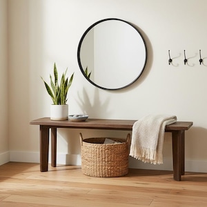 May include: A dark wood bench with four legs, a round black-framed mirror, and three black wall hooks. A white ceramic pot with a green snake plant, a small gray bowl, a woven seagrass basket, and a cream-colored textured throw blanket are included.