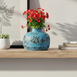 May include: A round, turquoise blue vase with a distressed, weathered finish containing a bouquet of vibrant red flowers with green leaves. The vase has a wide, bulbous body and a short, narrow neck, sitting on a light-colored wooden shelf.