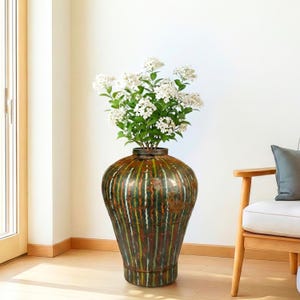 May include: A large, rounded metal floor vase featuring a vertical striped pattern in shades of dark green, copper, and white. The vase is filled with a bouquet of white flowers with green leaves, standing on a light-colored wooden floor.