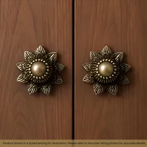 May include: Two identical drawer knobs shaped like multi-petaled flowers. Each knob features a smooth, matte gold-toned circular center surrounded by textured, antiqued brass-colored petals. The knobs are mounted on a vertical wood-grain surface.