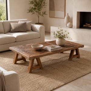 May include: A rectangular wooden coffee table with a weathered finish and A-frame legs. The tabletop features a carved pattern and holds a small ceramic bowl, a stack of books with the word Atlanta visible on the spine, and a round ceramic vase.
