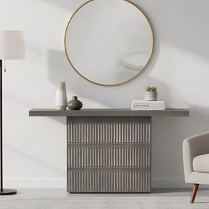 May include: A gray console table with a slatted base design, featuring a flat rectangular top. On the table sit a tall two-toned vase, a small dark gray rounded vase, a stack of three white books, and a small potted green succulent plant.