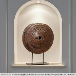 May include: A circular brown wooden shield featuring intricate carved geometric patterns and a central rectangular opening, mounted on a black metal stand with a rectangular base, displayed within a recessed arched wall niche.