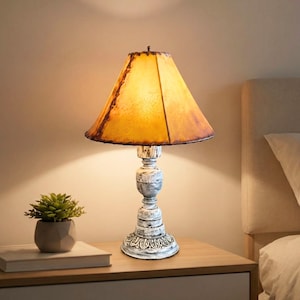 May include: A vintage-style table lamp with a distressed white base and a warm yellow lampshade. The lamp features a detailed, ornate design on the base and a textured lampshade with brown accents. A small potted plant and a book are on the table.