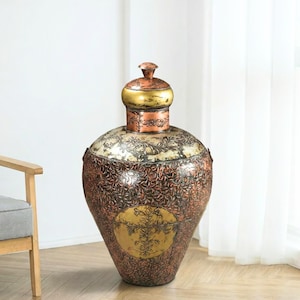 May include: A large, bulbous metal floor vase featuring a copper and dark bronze finish with intricate embossed floral patterns. The vase has a narrow neck, a rounded lid with a gold-toned top, and a prominent circular gold medallion on the front.