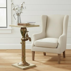 May include: A wingback chair with off-white fabric upholstery and wooden legs, positioned next to a side table featuring a square top and a base designed like a vintage metal water pump with a bronze finish, holding a pitcher and a small bowl.