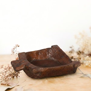May include: A rustic, dark brown wooden bowl with a square shape and a cross-like handle on one side. The bowl has a shallow interior and is set on a light-colored wooden surface. Dried flowers and leaves are in the background.