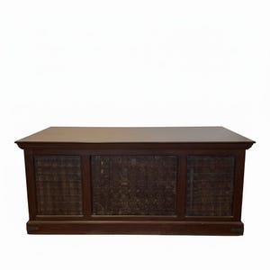 May include: A rectangular wooden desk with a dark brown finish. The front panel features three distinct sections, each containing a dark, textured grid pattern. The desk has a flat, smooth top surface and a solid base with small metal accents.