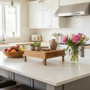 May include: A wooden riser with four turned legs sits on a white countertop. On the riser are two small potted succulents, a beige ceramic pitcher, and a stack of small plates. Nearby is a wooden bowl of fruit and a glass vase of pink peonies.