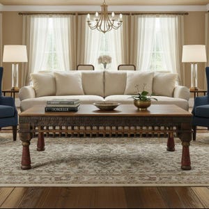 May include: A rectangular wooden coffee table with intricate carved detailing along the apron and four sturdy legs featuring red accents. The table surface holds two stacked books and a small brass bowl containing a green plant.