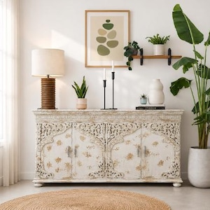 May include: A white wooden sideboard with intricate carved details and a distressed finish, featuring four cabinet doors. The sideboard is topped with a ribbed brown lamp, a potted snake plant, two black candle holders, and a white ceramic vase.