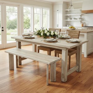 May include: A light-colored wooden dining table with a matching bench and woven chairs. The table is set with plates, silverware, and glasses. A floral arrangement sits in the center. The room has large windows and hardwood floors.