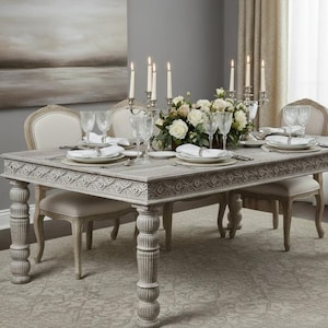 May include: A formal dining table set for a meal. The light gray table has carved detailing and is surrounded by matching chairs. The table is set with white plates, silverware, and wine glasses. A floral centerpiece and lit candles complete the elegant setting.