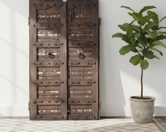 May include: A pair of tall, weathered dark brown wooden doors featuring horizontal metal straps, decorative metal studs, and circular iron ring handles. The doors have a distressed finish with visible remnants of white paint in the recessed panels.