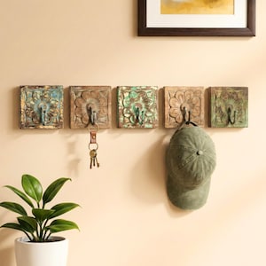 May include: Five square wooden wall hooks with floral carvings and metal hooks. The hooks are painted in shades of blue, green, and brown. A green cap and a set of keys hang from the hooks. A potted plant is in the foreground.