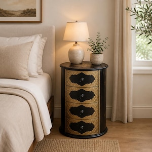 May include: A round black bedside table with four drawers, each featuring ornate gold-colored filigree detailing and a small black knob. The table is topped with a cream-colored ceramic table lamp and a small potted green plant.
