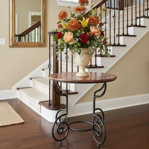 May include: A round side table with a hammered copper-toned top and a black metal base featuring scrolled legs. A cream-colored decorative urn sits on the table, filled with a large arrangement of orange, red, and yellow flowers and green foliage.