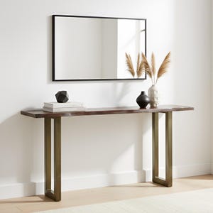 May include: A rectangular console table with a dark brown live-edge wooden top and two brushed brass U-shaped legs. On the table are two stacked white books, a black geometric object, a black vase, and a white vase with dried pampas grass.
