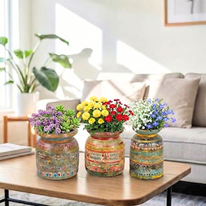 May include: Three decorative, hand-painted ceramic vases filled with colorful flowers and succulents. The vases have a rustic, aged appearance with intricate patterns in shades of blue, green, red, and yellow. The vases are displayed on a wooden table.