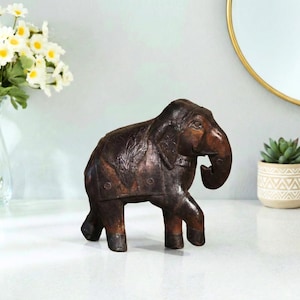 May include: A dark brown leather elephant figurine featuring intricate embossed patterns on its back and sides. The elephant is depicted in a walking stance with its trunk curved downward, showcasing detailed stitching and a smooth, polished finish.