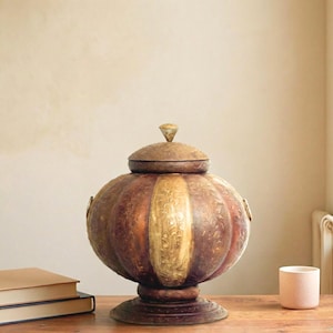 May include: A round, lidded metal vessel featuring alternating vertical panels of burnished copper and polished gold tones. The surface is decorated with intricate floral engravings, and the container rests on a circular pedestal base.