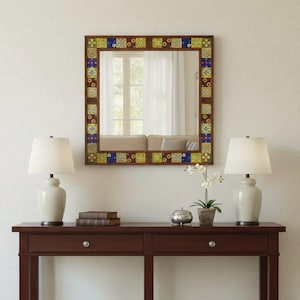 May include: A square wall mirror featuring a dark wood frame decorated with a border of colorful, patterned tiles in shades of blue, yellow, and green. The mirror reflects a window with white curtains and a portion of a sofa.